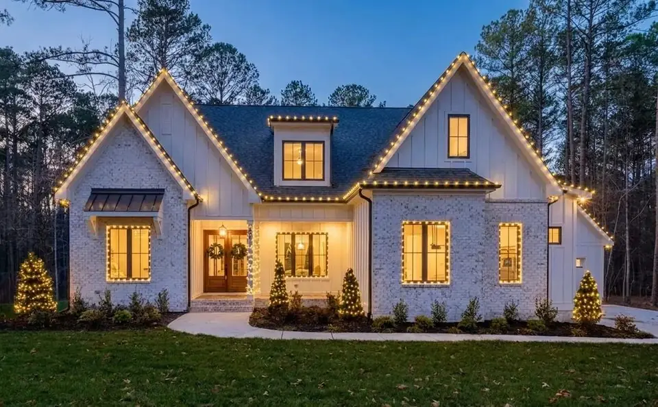 The same house at dusk with warm white lights along every roofline, window and shrub